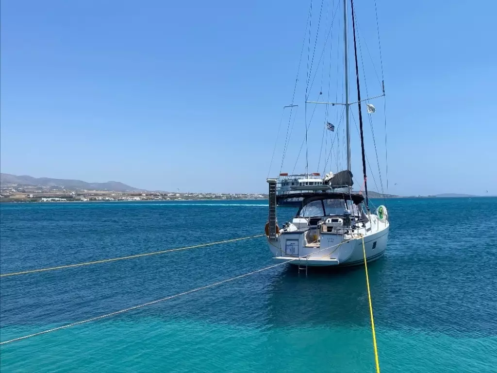 A sailboat anchored in clear blue water, with ropes extending from the boat to the shore in the background under a clear blue sky.